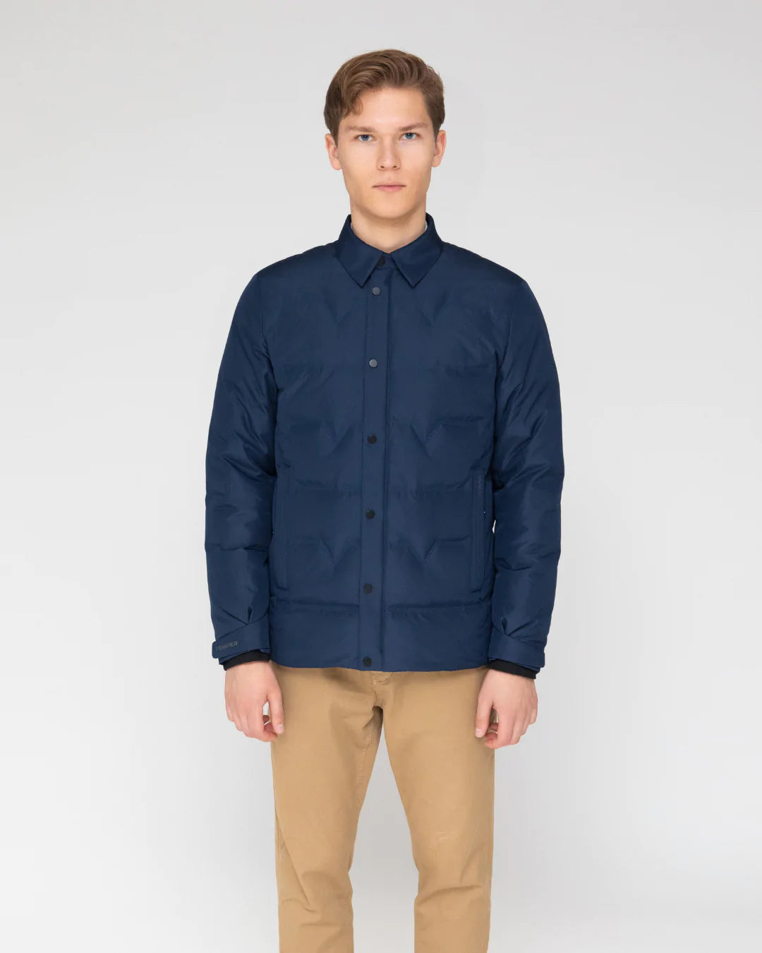 A man wearing a navy blue and black puffer jacket stands outdoors with his hands in his pockets. The jacket is hip-length with a stand collar and elasticized cuffs. It appears well-insulated and suitable for winter weather. The Active Puffer Jacket has a sporty yet refined look, seeming functional for everyday wear. The premium, waterproof and windproof fabric would make it an ideal semi-casual travel piece able to withstand weather conditions. This versatile jacket works for various situations.