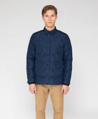 A man wearing a navy blue and black puffer jacket stands outdoors with his hands in his pockets. The jacket is hip-length with a stand collar and elasticized cuffs. It appears well-insulated and suitable for winter weather. The Active Puffer Jacket has a sporty yet refined look, seeming functional for everyday wear. The premium, waterproof and windproof fabric would make it an ideal semi-casual travel piece able to withstand weather conditions. This versatile jacket works for various situations.